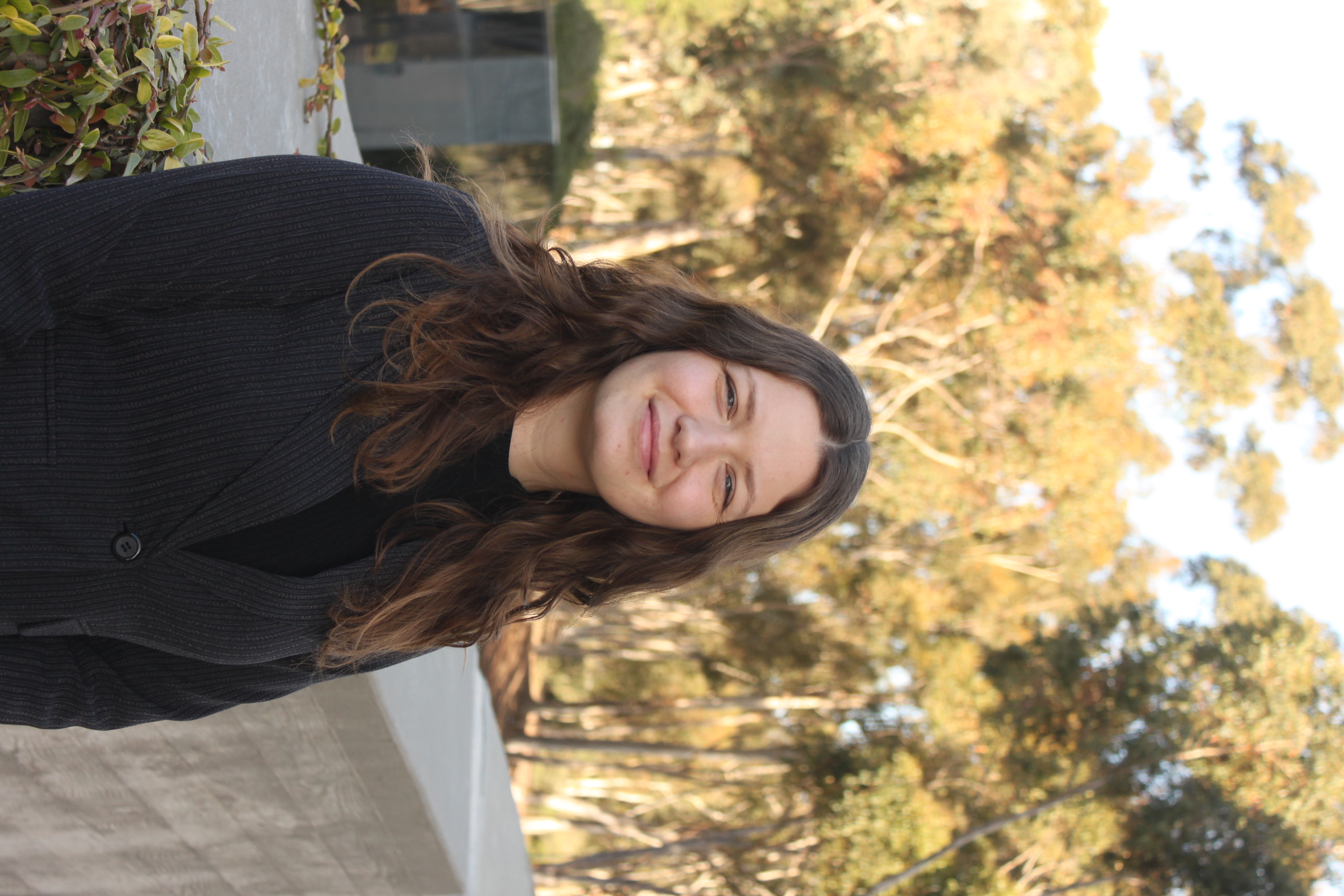Woman with long brown hair smiling and standing in front of a brick wall with a black jacket and black shirt